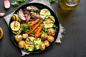 Image of a black plate sitting on a white cloth towel with frayed edges.  In the corner are two pieces of garlic and parsley.  On the plate are roasted vegetables: zucchini, new potatoes, and red onions around the outside with carrots sliced lenthwise in 